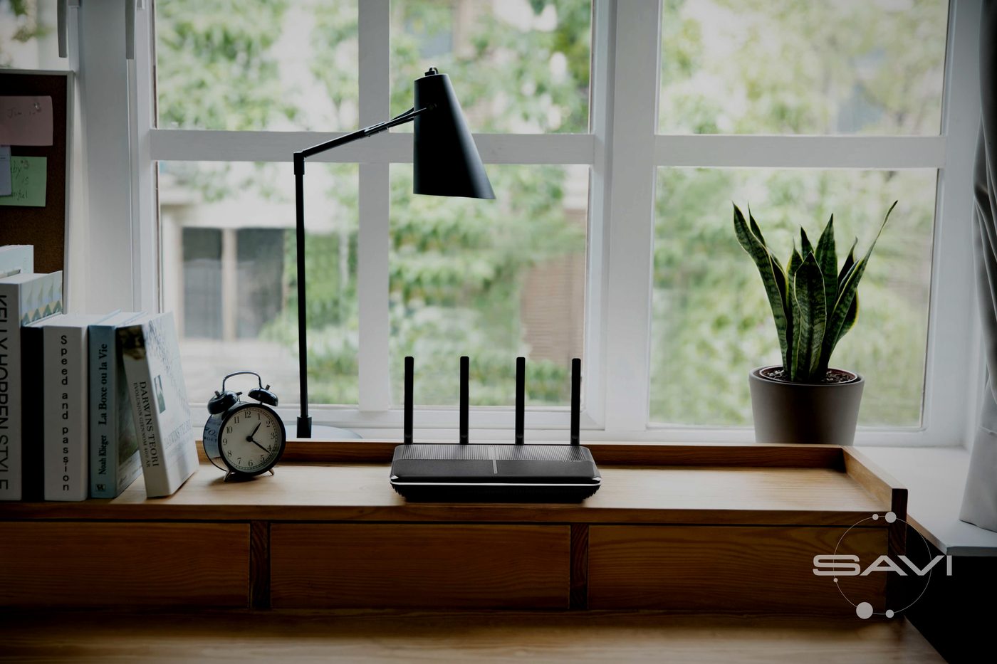 Чёрный wifi-роутер с антеннами стоит на деревянной тумбе у окна в комнате, рядом настольная лампа, будильник и комнатное растение